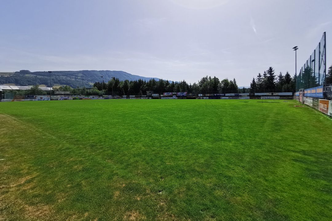 Fu&szlig;balltrainingsplatz im Salzkammergut mit gr&uuml;nem Rasen, Tor, Spielern, blauer strahlender Himmel, umgeben von Berglandschaft.
