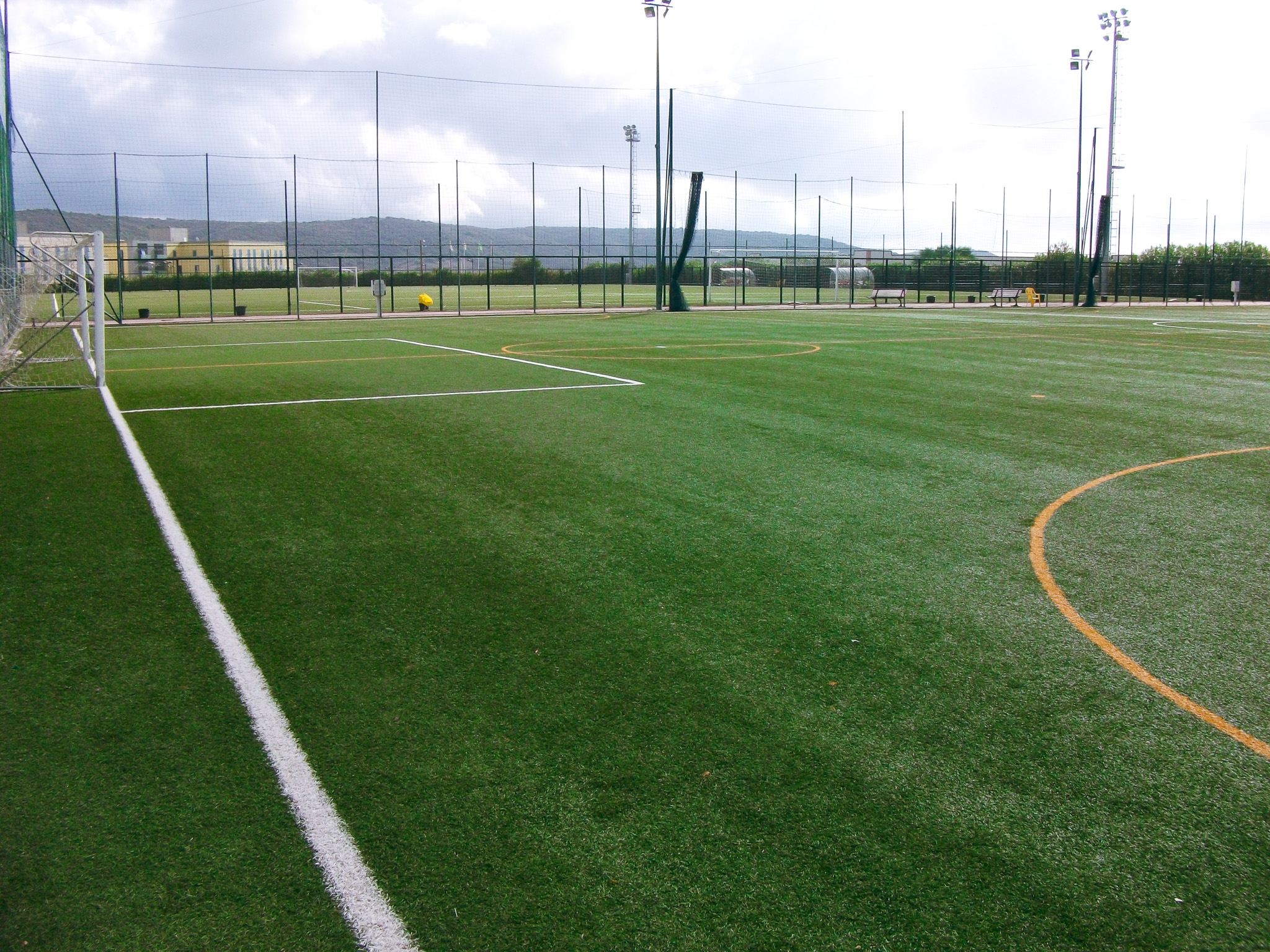 Fu&szlig;balltrainingsplatz auf Sardinien mit gr&uuml;nem Kunstrasen, Tor, B&auml;nken und Flutlichtmasten, umgeben von Netzen und Berglandschaft.