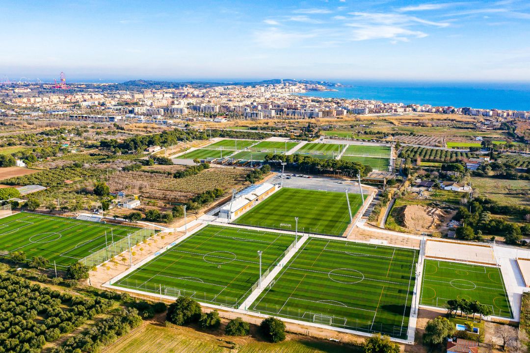 Luftaufnahme einer modernen Fu&szlig;balltrainingsanlage in Spanien mit mehreren gepflegten Kunstrasen- und Naturrasenpl&auml;tzen, umgeben von Olivenhainen und landwirtschaftlichen Fl&auml;chen, mit Blick auf eine K&uuml;stenstadt und das Mittelmeer im Hintergrund.