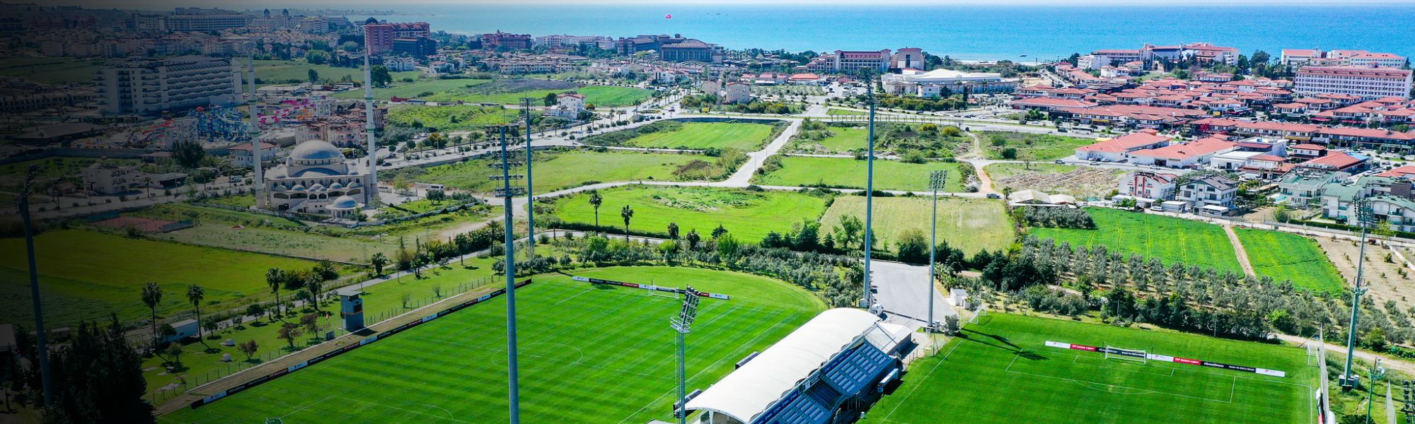 Luftaufnahme eines Fu&szlig;balltrainingslagers in der T&uuml;rkei mit zwei Rasenpl&auml;tzen, Trib&uuml;ne und Blick auf die Stadt und das Meer.