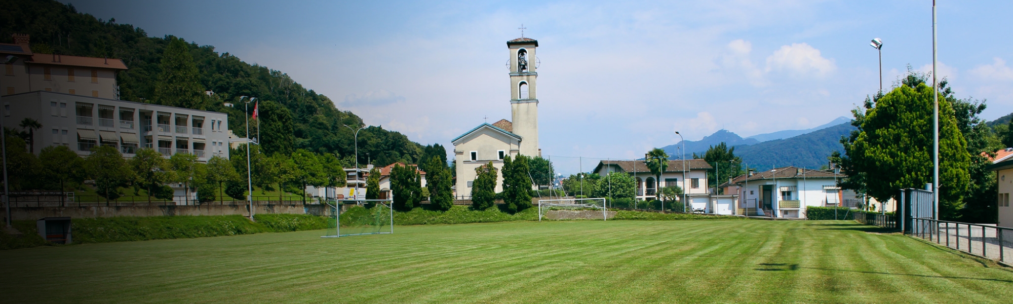 Panoramaaufnahme eines Fu&szlig;ballfeldes im Tessin, eingebettet in eine gr&uuml;ne Landschaft mit einer Kirche und typisch italienischen H&auml;usern im Hintergrund. Perfekte Bedingungen f&uuml;r ein Fu&szlig;balltrainingslager in der Schweiz.