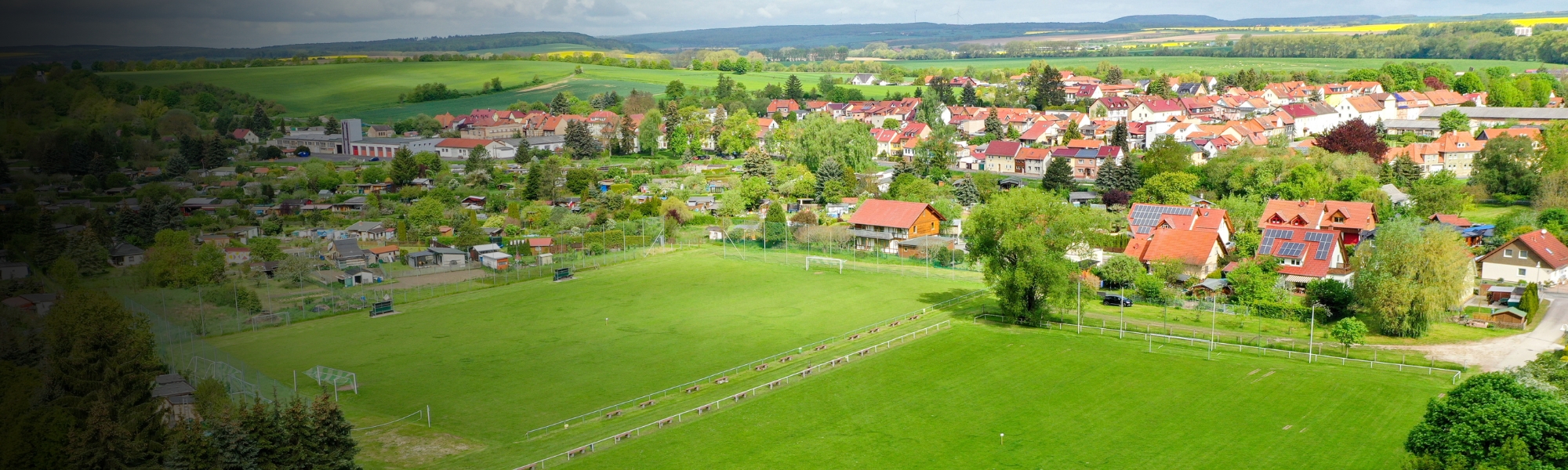 Fu&szlig;ballplatz in l&auml;ndlicher Umgebung in Th&uuml;ringen, umgeben von gr&uuml;nen Wiesen, Kleing&auml;rten und einer Dorfkulisse mit roten D&auml;chern.