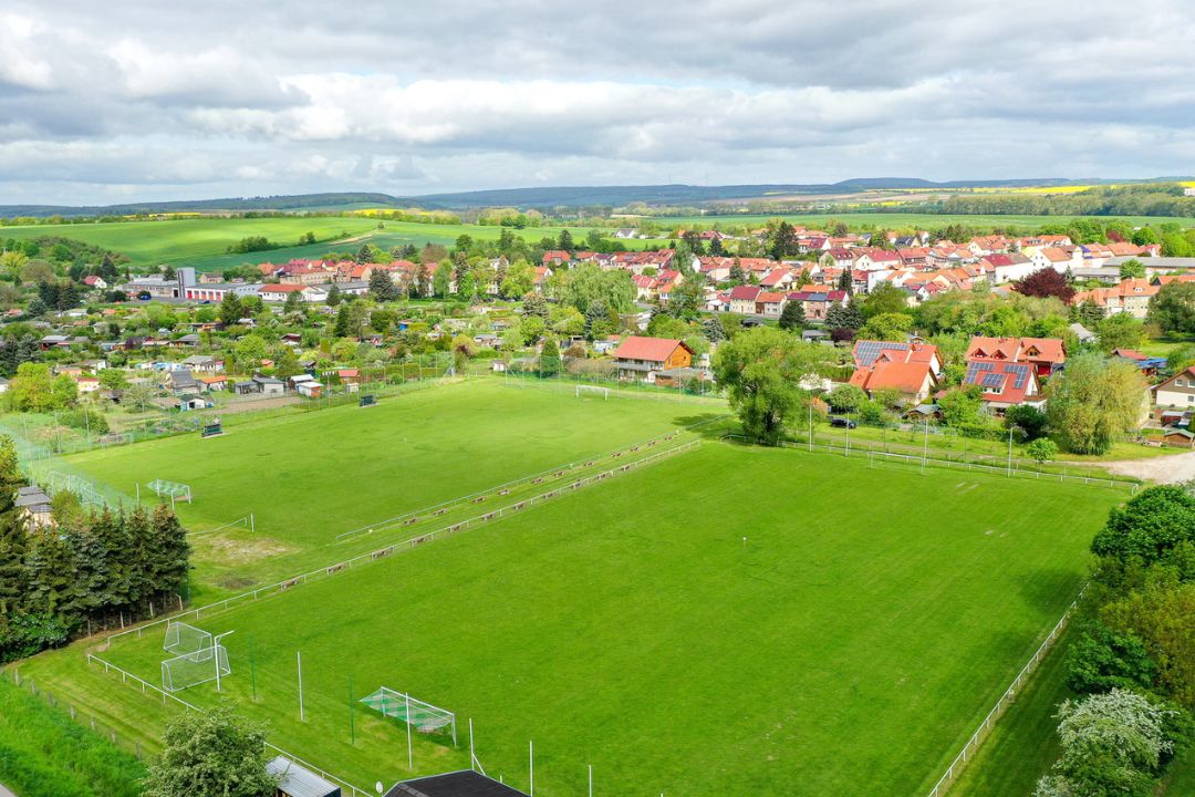 Luftaufnahme eines Fu&szlig;ballplatzes in Th&uuml;ringen, eingebettet in eine idyllische Landschaft mit Feldern, B&auml;umen und einer kleinen Wohnsiedlung im Hintergrund.