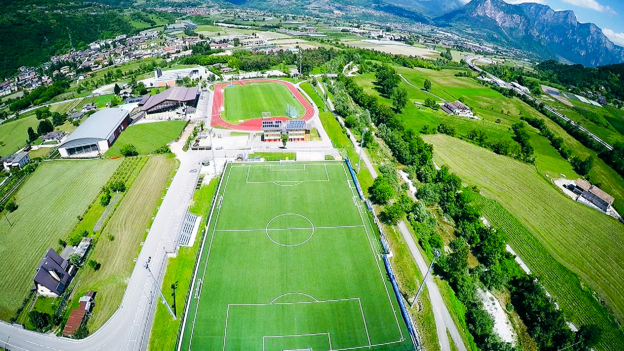 Sportanlage in Trentino mit Fu&szlig;ballplatz, Leichtathletikstadion, Sporthalle und gr&uuml;nen Feldern, eingebettet in eine idyllische Berglandschaft.