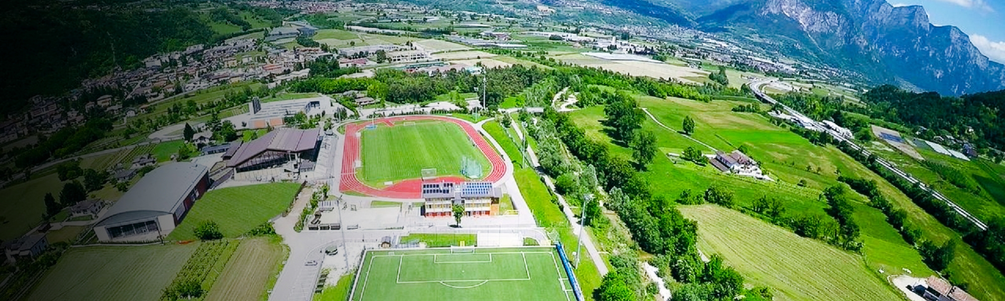 Luftaufnahme eines modernen Fu&szlig;ballplatzes mit Trib&uuml;ne und angrenzender Leichtathletikanlage inmitten der gr&uuml;nen Landschaft von Trentino, umgeben von Bergen und kleinen Ortschaften.