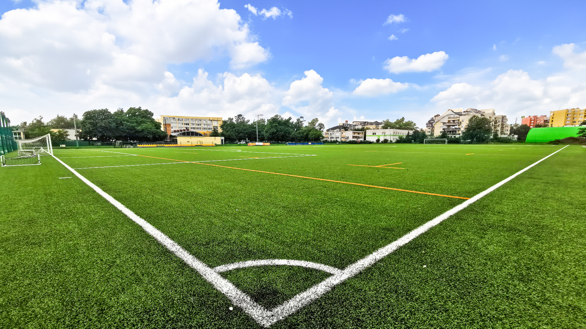 Moderner Kunstrasenplatz mit klaren Spielfeldmarkierungen, umgeben von Wohngeb&auml;uden und Sporteinrichtungen.