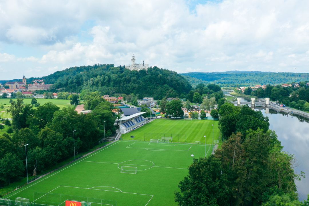 Fu&szlig;ballplatz in Tschechien, umgeben von Wald und H&uuml;geln, mit Blick auf ein Schloss und eine Stadt.