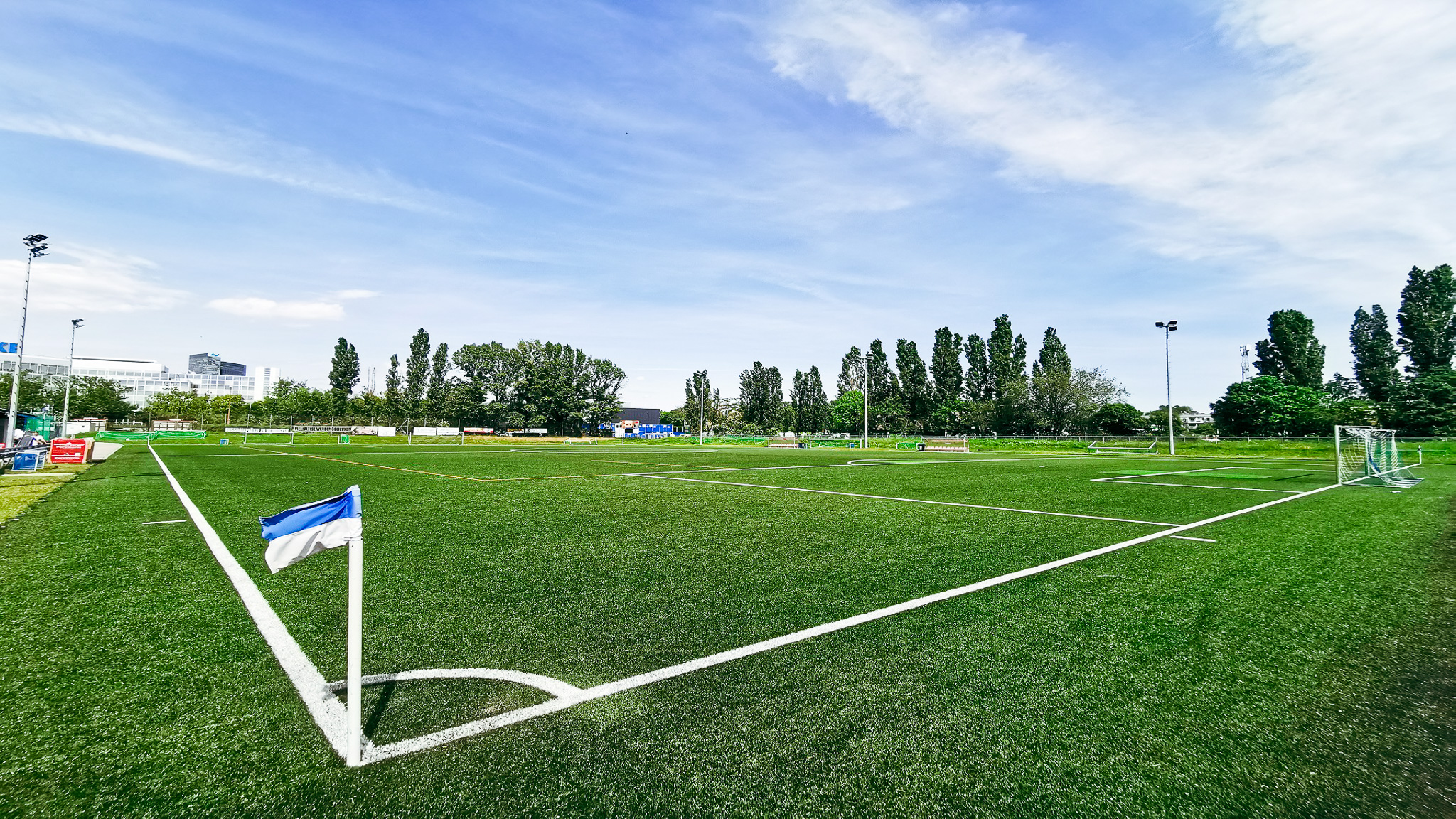 Fu&szlig;ballplatz mit Kunstrasen und blauer Eckfahne in Wien, umgeben von B&auml;umen und modernen Geb&auml;uden im Hintergrund.