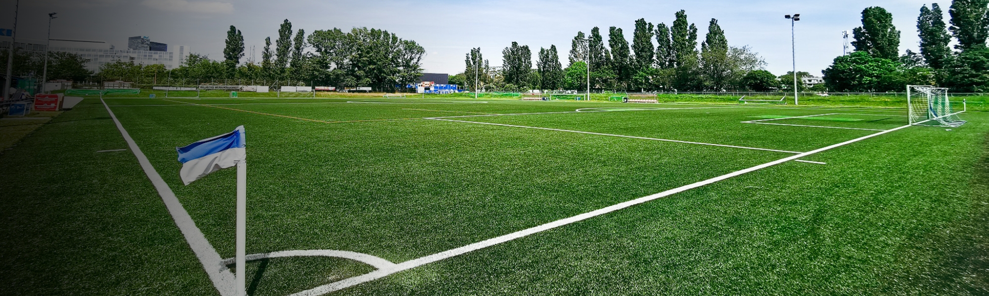 Fu&szlig;ballplatz mit Kunstrasen und blauer Eckfahne in Wien, umgeben von B&auml;umen und modernen Geb&auml;uden im Hintergrund.
