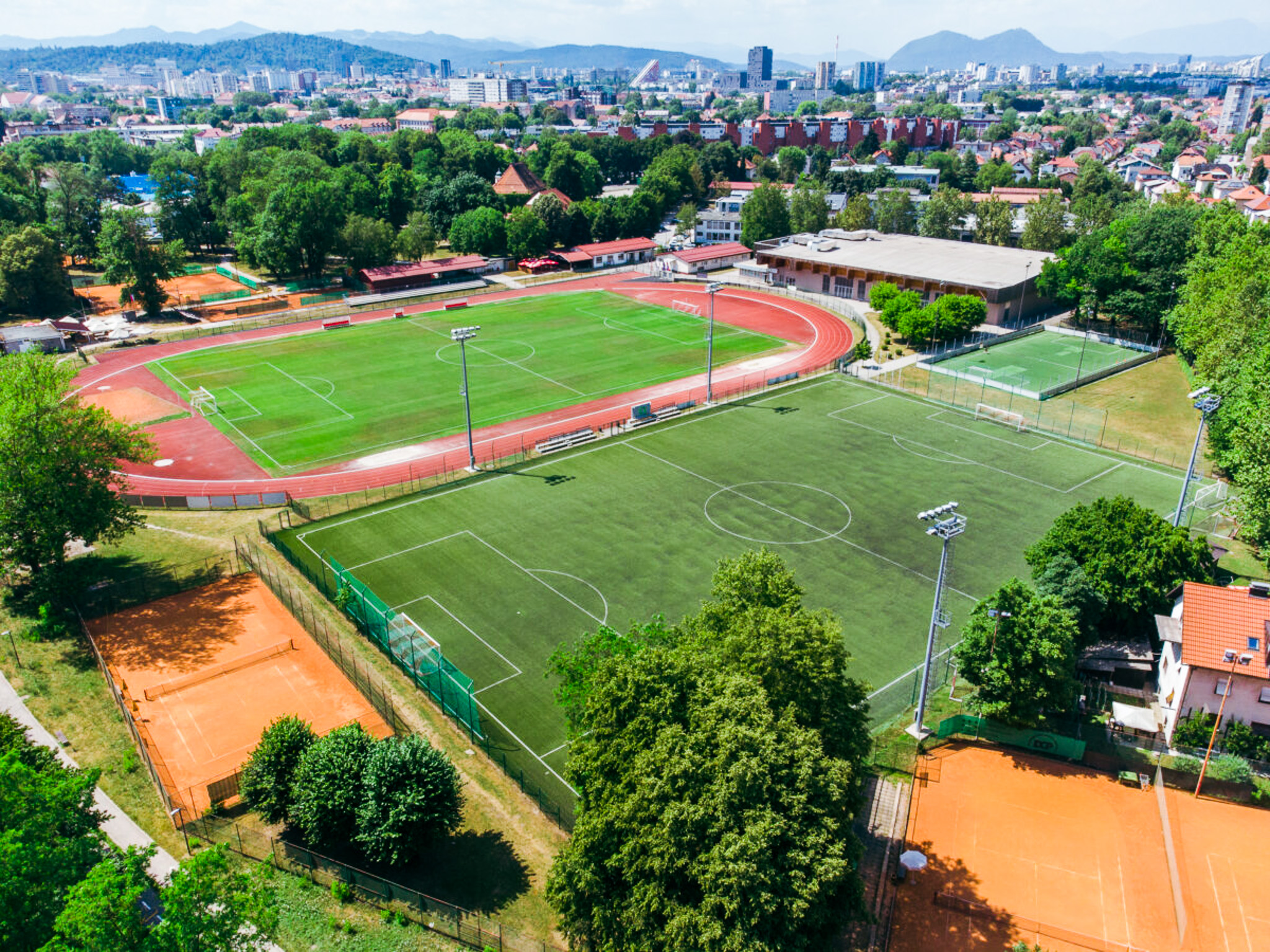Fu&szlig;balltrainingslager mit mehreren Spielfeldern, Laufbahn und Tennispl&auml;tzen, eingebettet in eine st&auml;dtische Umgebung mit Bergen im Hintergrund.