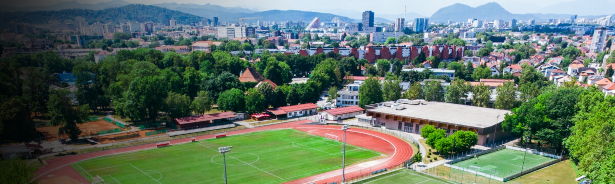 Fu&szlig;balltrainingslager mit mehreren Spielfeldern und Laufbahn, eingebettet in eine st&auml;dtische Umgebung mit Bergen im Hintergrund.