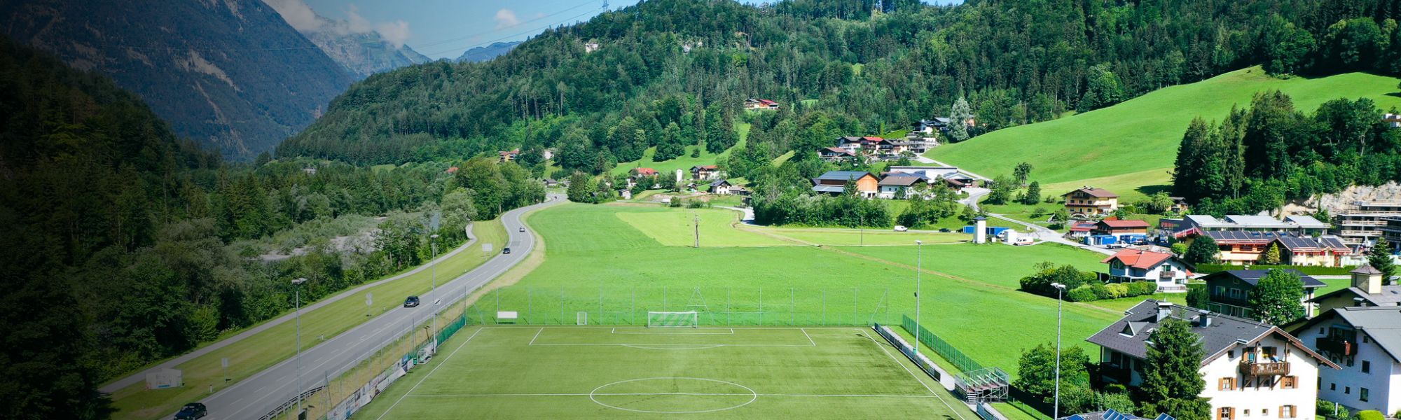 Panoramaansicht eines Fußballplatzes mit einem kleinen Dorf und Bergen in Österreich im Hintergrund.
