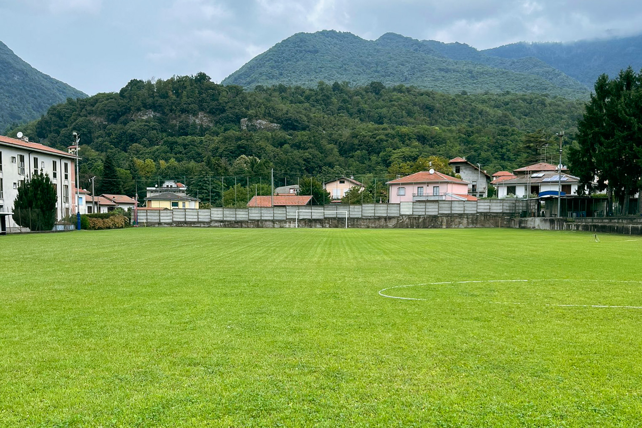 Fu&szlig;balltrainingsplatz auf Sardinien mit gr&uuml;nem Kunstrasen, Tor, B&auml;nken und Flutlichtmasten, umgeben von Netzen und Berglandschaft.